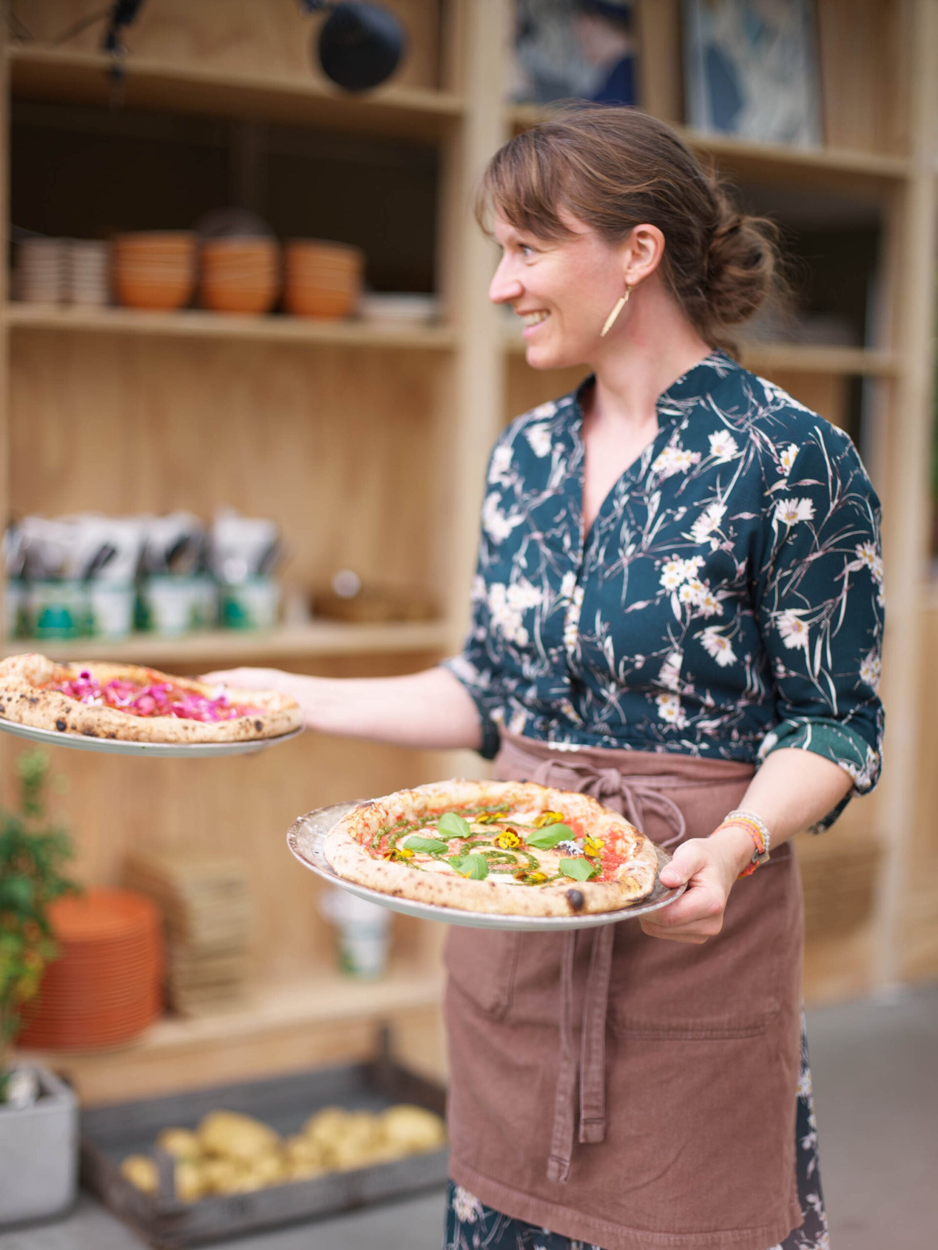 Kvinne med blomstrete skjorte og brun forkle, som bærer to pizzaer med grønnsaker og urter.