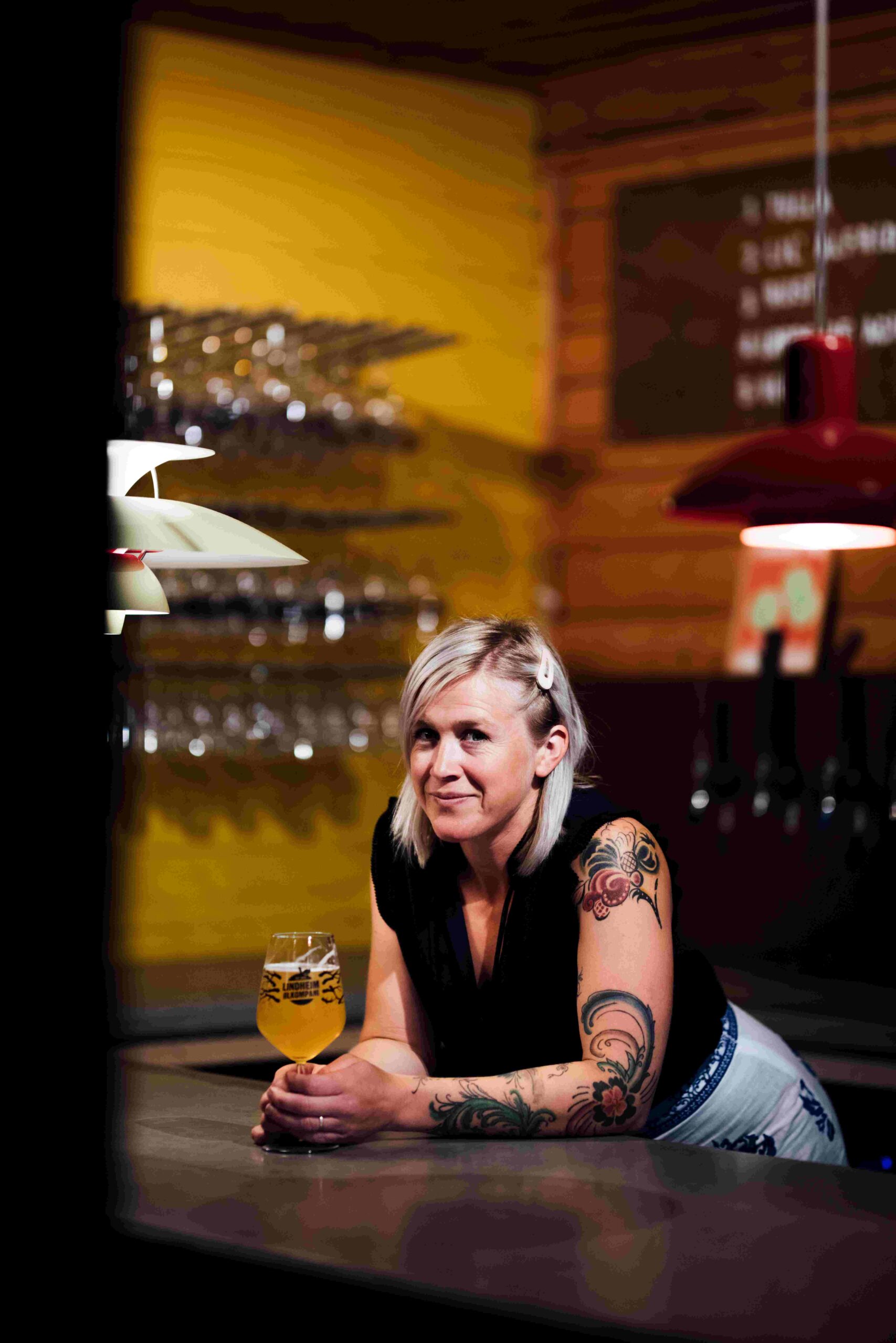 Bartender med tatoveringer, smilende ved baren med et glass øl i hånden. Fargerik bakgrunn i baren.