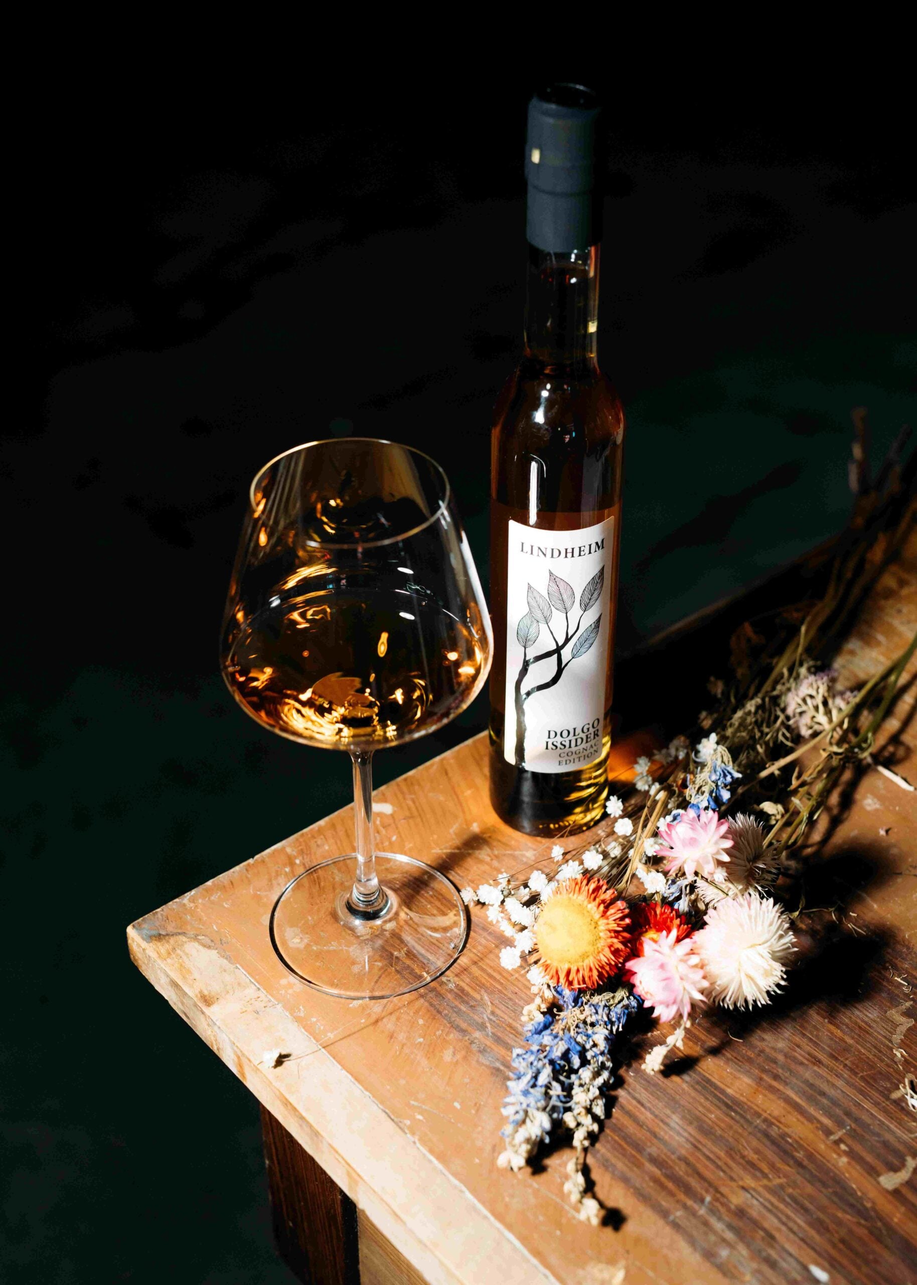 Flaske med Lindheim cider ved siden av et glass, dekorert med tørkede blomster på et trebord.