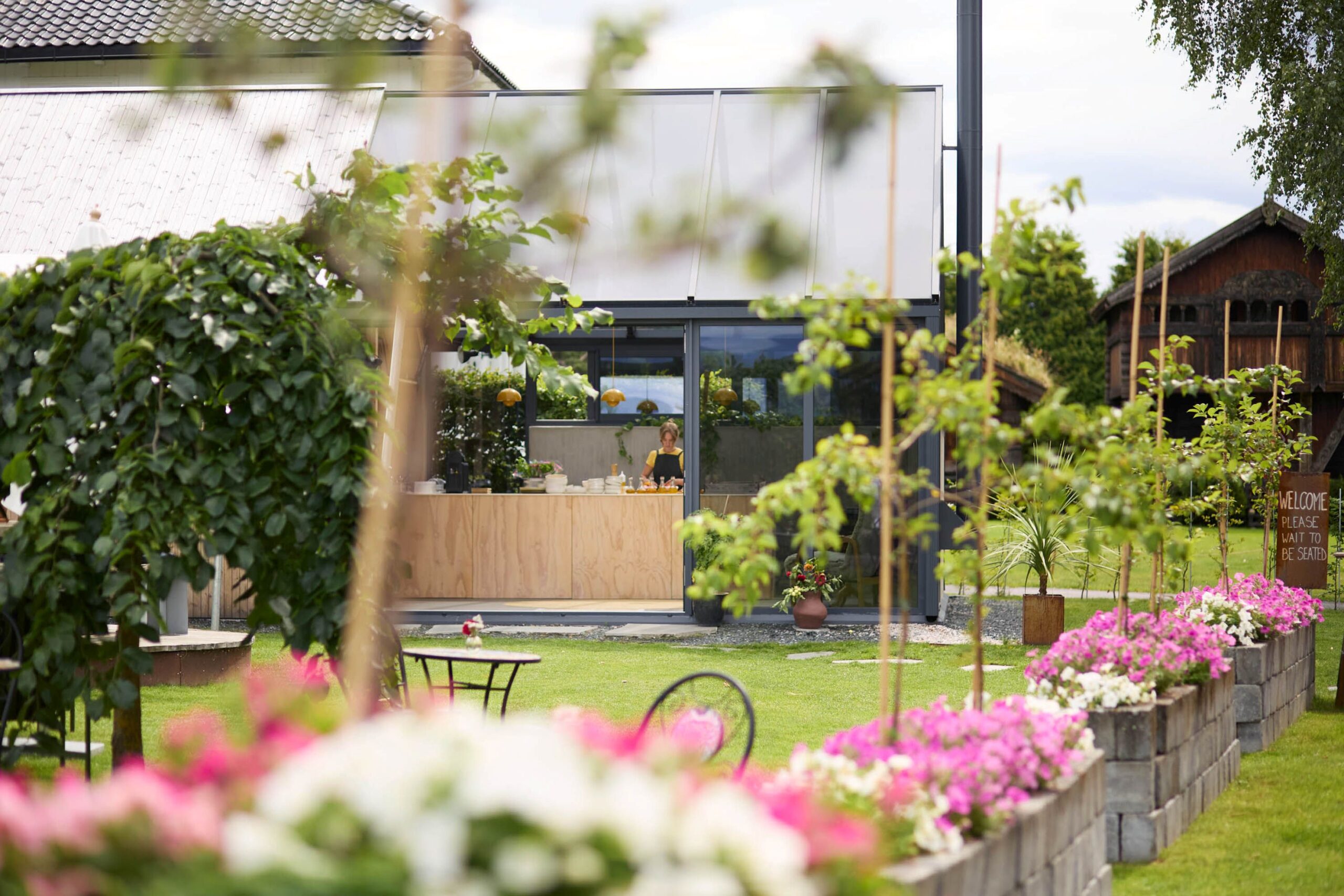 Kafe med hage, blomsterbed og servitør som jobber i en koselig uteservering. Solfylt dag med frodig grønt.