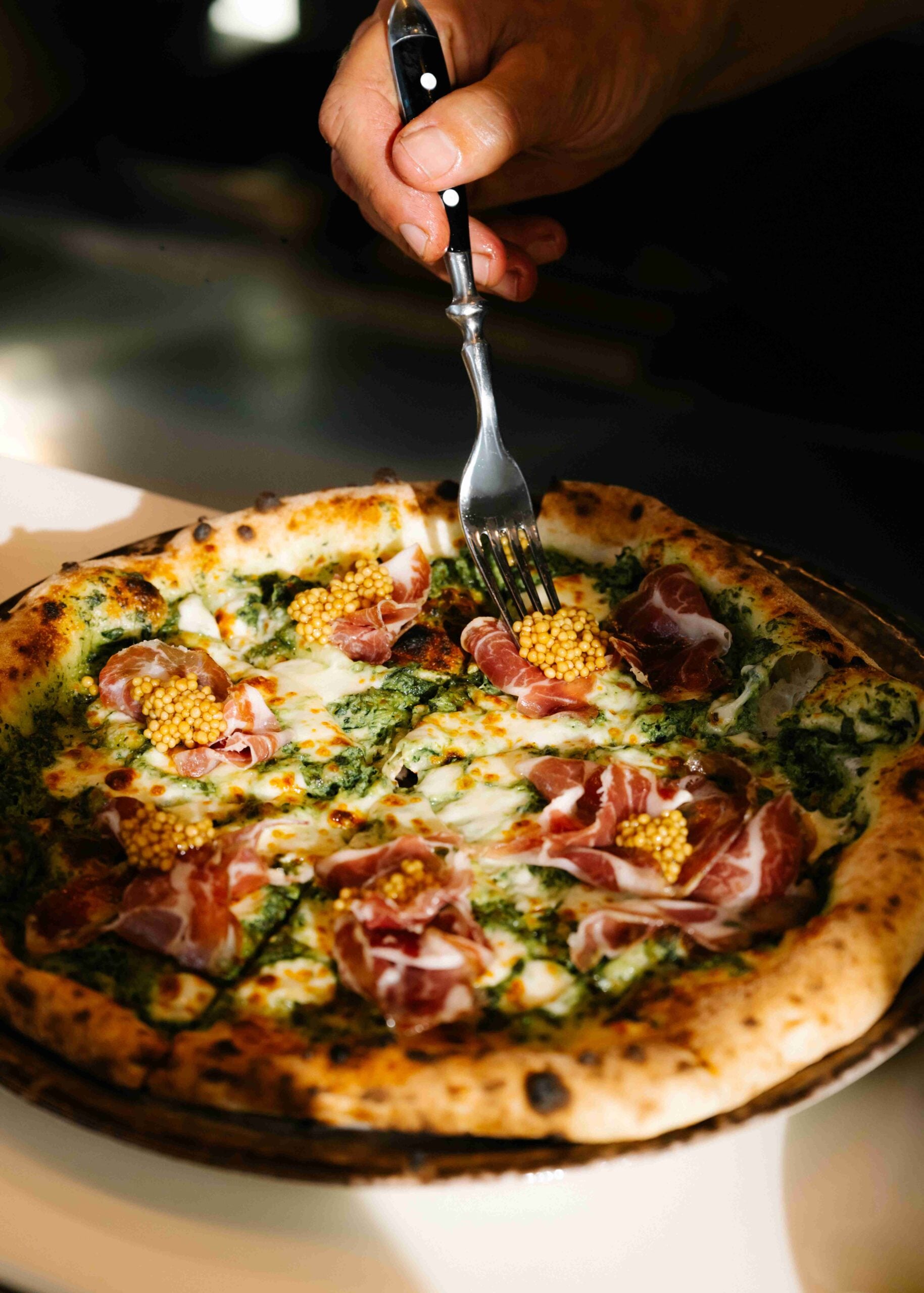 Hånd med gaffel som skjærer i en pizza med spekeskinke og sennepsfrø, ser smakfull ut. Grønn pesto på bunnen.